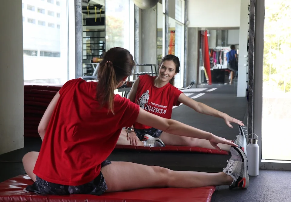 A woman in a red Crossjoin t-shirt sits on a gym mat stretching her leg while looking at her smiling reflection in a large wall mirror.